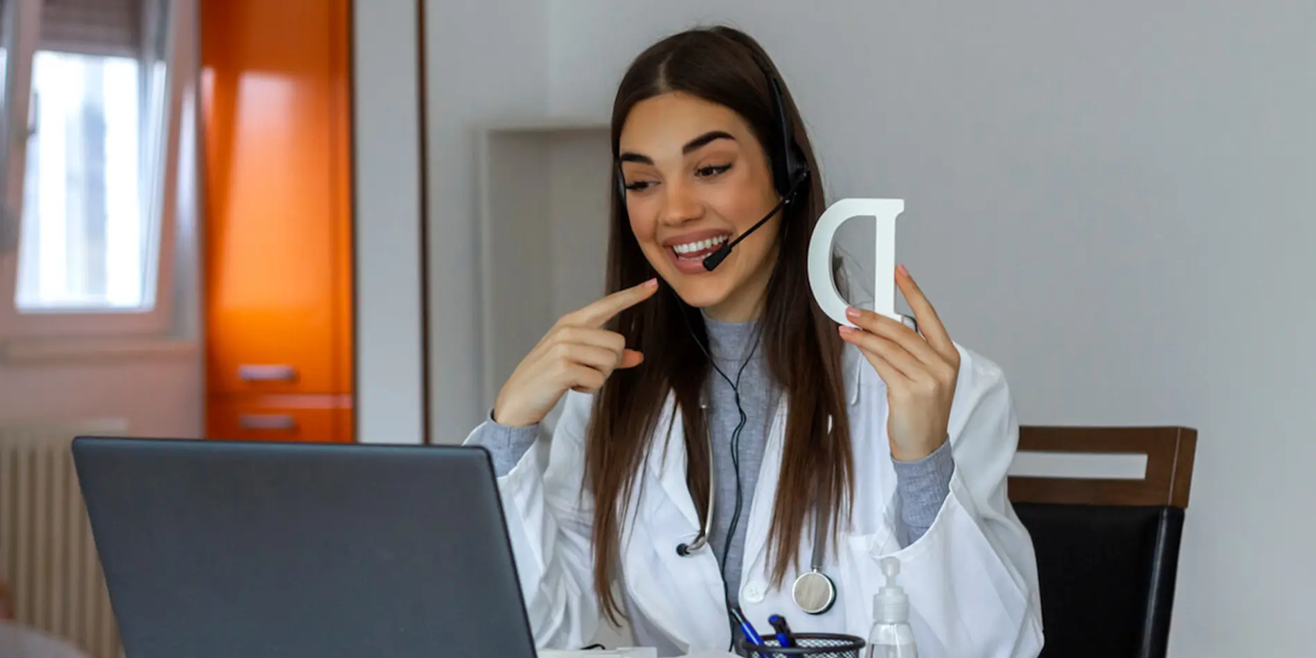 A person wearing a lab coat holds up a wooden letter "D" to an audience streaming on the laptop while wearing a headset.