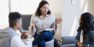 A person is speaking with two youths on their left and right. They are in a white room sitting on sofas.