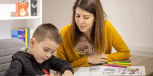 A teacher is looking over a student's writing. Both are sitting at a desk in a classroom. There are coloring pens and completed drawings on the table.