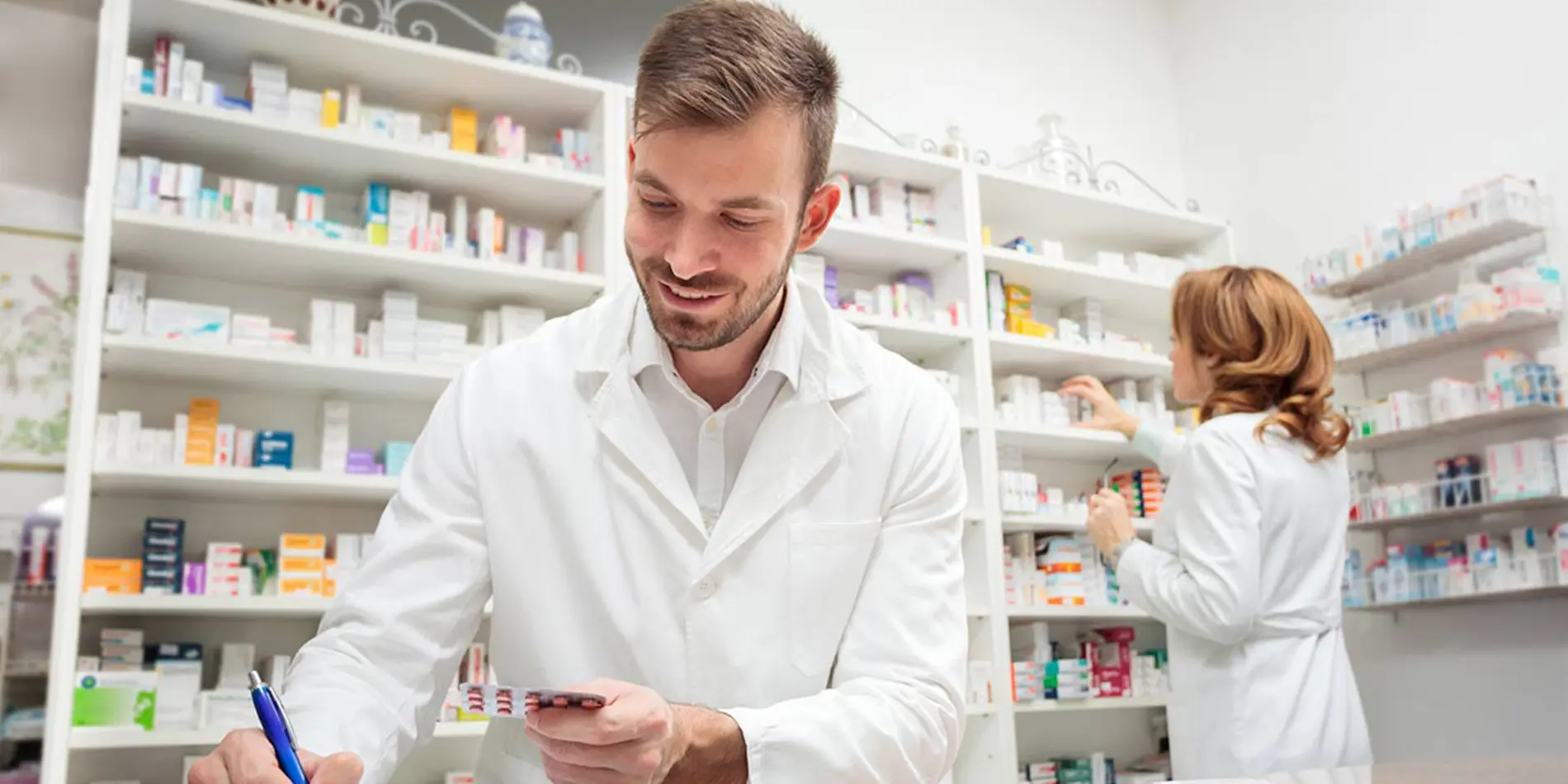 A pharmacist is looking down and writing as they hold a sheet of tablet pills. Pills line the wall in the room. Another pharmacist is in the background, facing the wall, organizing.