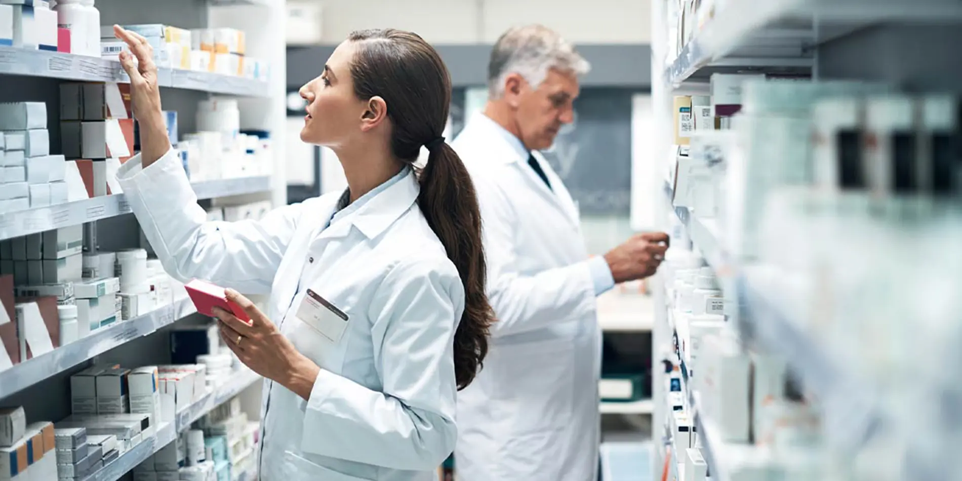 Two people are looking at pill bottles on shelves in a pharmacy. They are wearing lab coats.