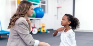 A person wearing business professional attire holds their hand to their throat. A young person, in front of them, mimics the action. They are in a gym setting.