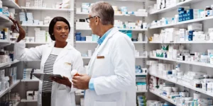 Two people in white lab coats inside a pharmacy. Medicine bottles line the wall.