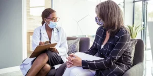 Two people are looking at each other, wearing masks. They sit on a sofa. One is wearing a lab coat, a stethoscope, and is holding a clipboard taking notes. The other person has clasped hands around their crossed legs. A window is behind them.