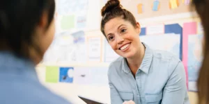 A person is smiling at someone whom is shown from the back on the left side of the shot. They are in a classroom.