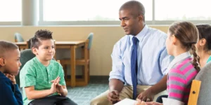 A teacher is sitting in a circle with children around them in chairs.