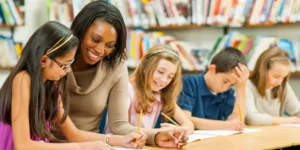 Four students sit at a table and are writing. There is a teacher in between them, smiling. They are in a library, books are behind them.