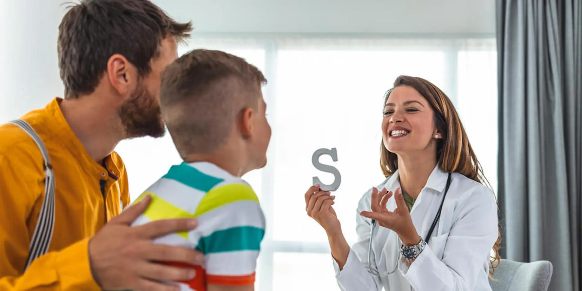 A speech language pathologist is pronouncing the letter "S" while also holding up a wooden "S" to a young person and their guardian.