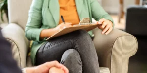 A person is shown with their legs crossed and a clipboard. They are writing and sitting in a sofa. There is a person shown across from them, only their clasped hands shown.