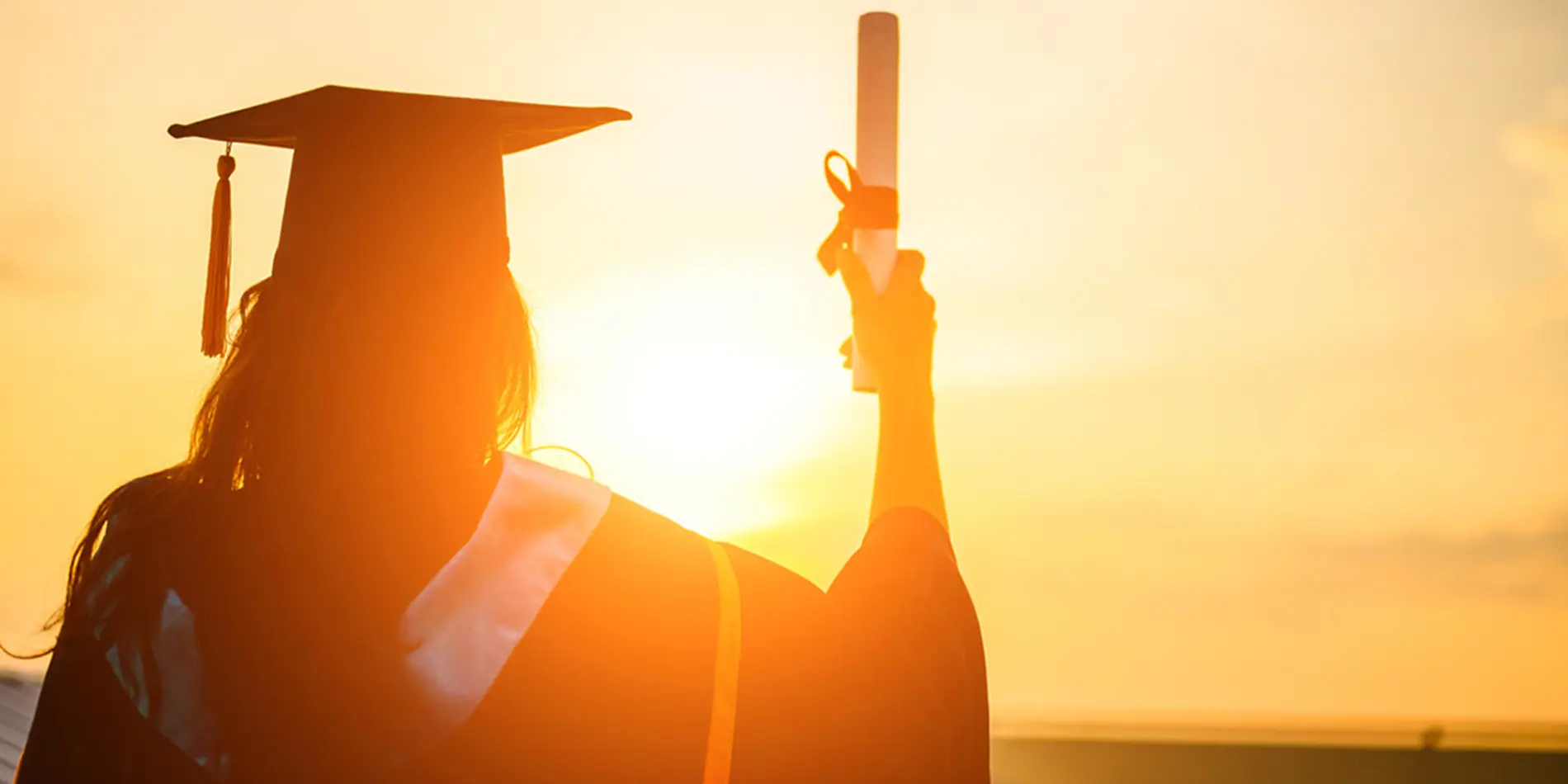 A person with traditional graduation regalia-- like a cap with tassel, hood, and robe-- is holding a graduation scroll facing a sunset shown from the back.