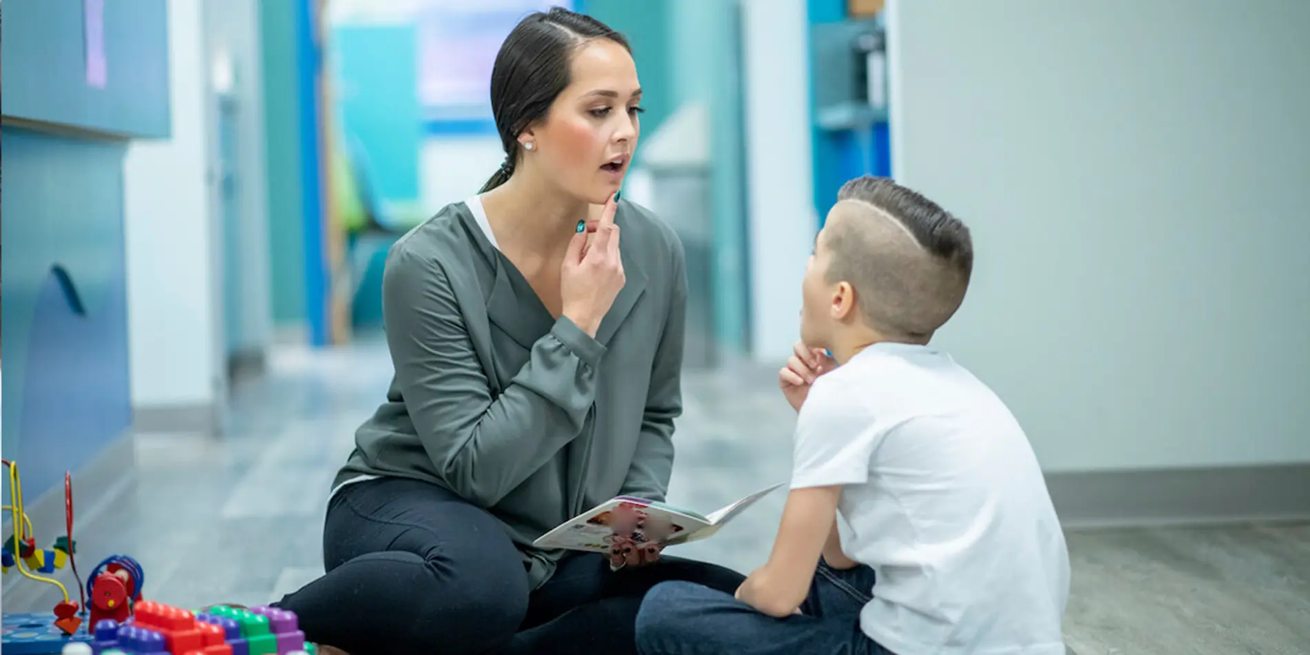A speech-language pathologist is sitting on the ground in a classroom and pointing to her mouth as she pronounces something. A young person sits across from her and is mimicking the expression.