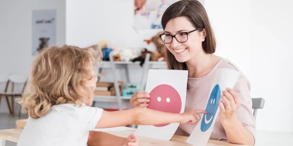 A teacher is holding up two sheets of paper: one with a blue smiley face, the other with a pink one. A child is sitting across from the teacher pointing at the blue one. They are in a white room.