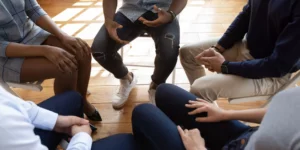 A group of people with varying skin tones sit in a circle on folding chairs. Only their laps are shown.