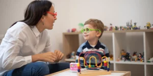 A person is looking to the right at a young person. They are looking at each other and wearing colorful play glasses. Developmental toys are on the table in front of them and in the shelves behind them.