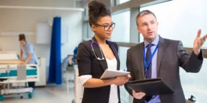 Two people with clipboards are talking to each other in a hospital room. They are dressed in business professional attire.