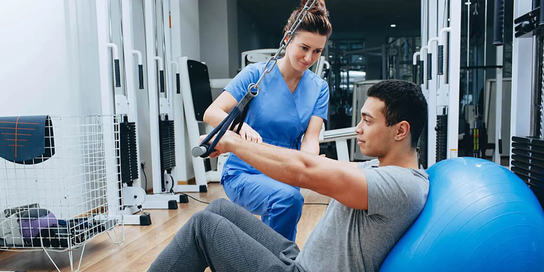 Two people are in a gym. One is doing exercises on an exercise ball. The other is wearing scrubs and aiding in the exercise.