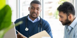 Two men look at a piece of paper in a folder. One is wearing business professional attire and a stethoscope.