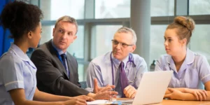 Four people are sitting around a table with a laptop. Two are wearing a nurse's uniform, the other two are wearing business professional attire.