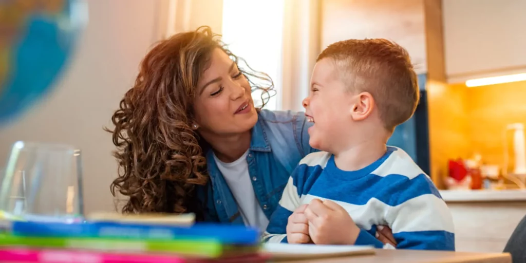 A person is looking at a laughing young person while sitting at a table. There are developmental toys in front of them.