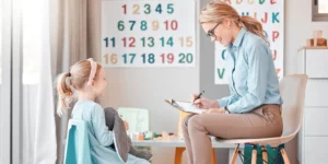 A person is sitting, smiling, and looking down at their clipboard while a young person across from them speaks, also sitting. They are in a classroom setting with numbers and letters on the wall.