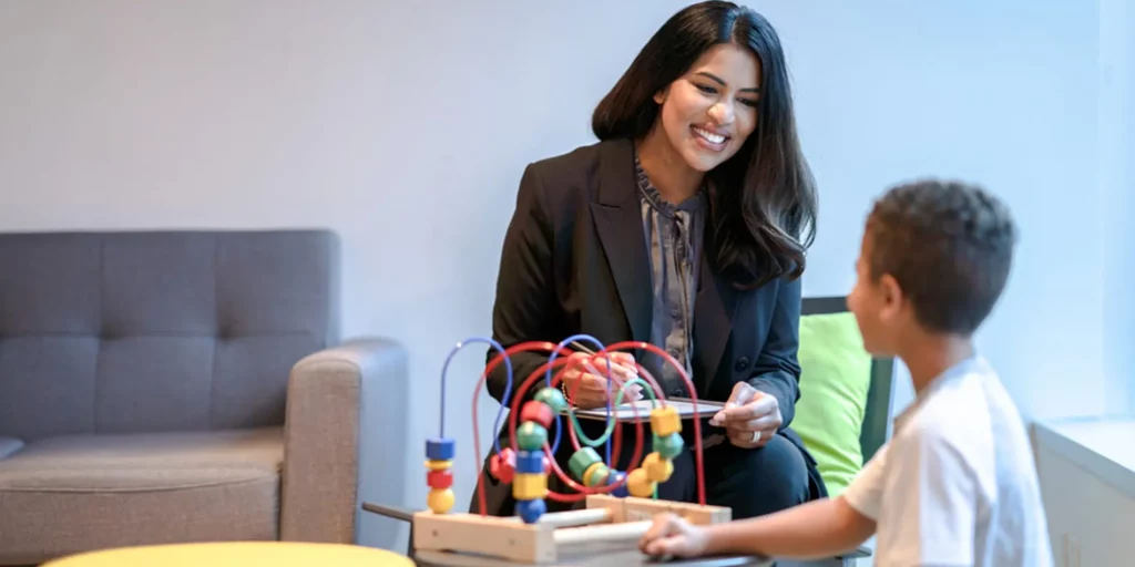 A person writing on a clipboard is smiling at a young person playing with a developmental toy.