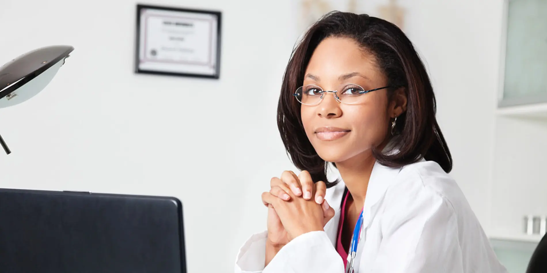 A woman wearing glasses sitting in front of a laptop computer wearing a lab coat and stethoscope. Her medical degree is hung up on the wall behind her.