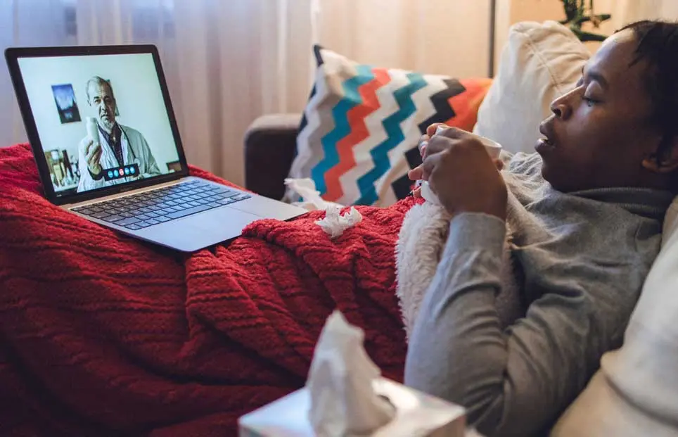 Woman of color in bed nursing a cold and speaking to her doctor on her computer