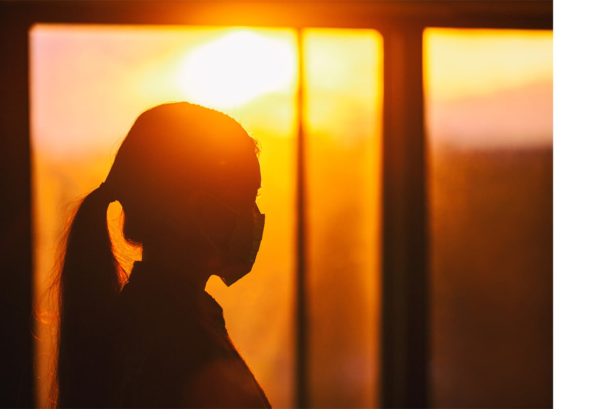 Woman in a mask looking out a window at a sunset