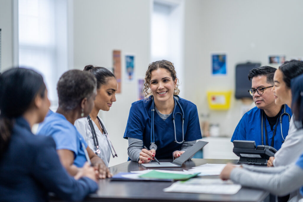 A small group of medical professionals gather around a table as they meet together to discuss patient cases. They are each dressed professionally and have files scattered between them for reference.