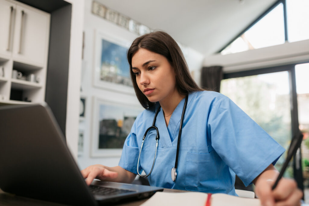 Nursing student in uniform, working from home and checking and advising her patient using a laptop to talk to them remotely, writing some notes