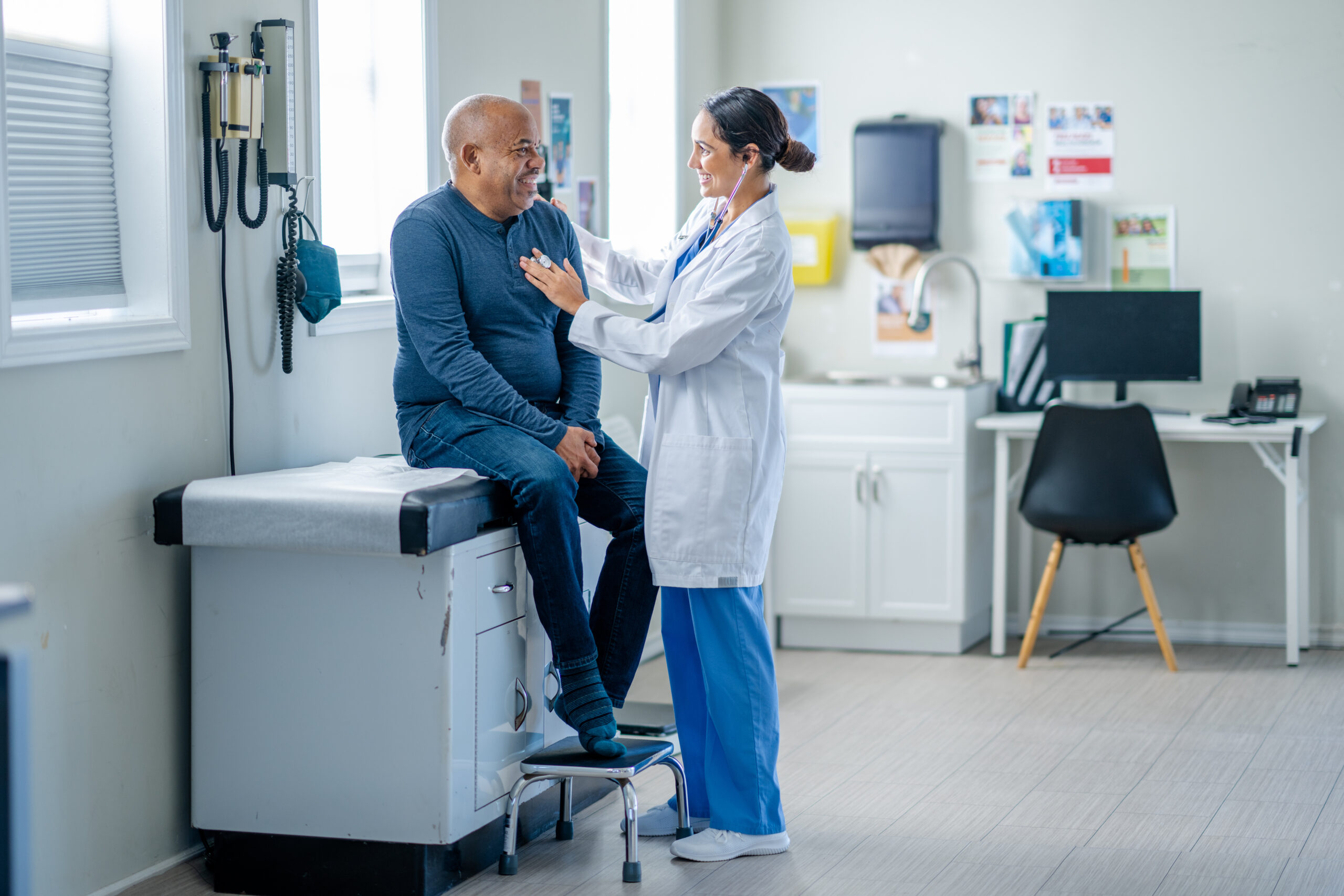 A senior gentleman of African decent sits up on an exam table during a routine check-up. He is sitting still and quiet as his female doctor listens to his heart.