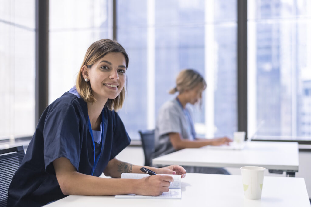 Portrait of a female nurse in a modern classroom environment. Wearing medical scrubs and writing notes in a notebook. The student is looking directly at the camera and smiling. Another female classmate can be seen in the background working on a laptop.