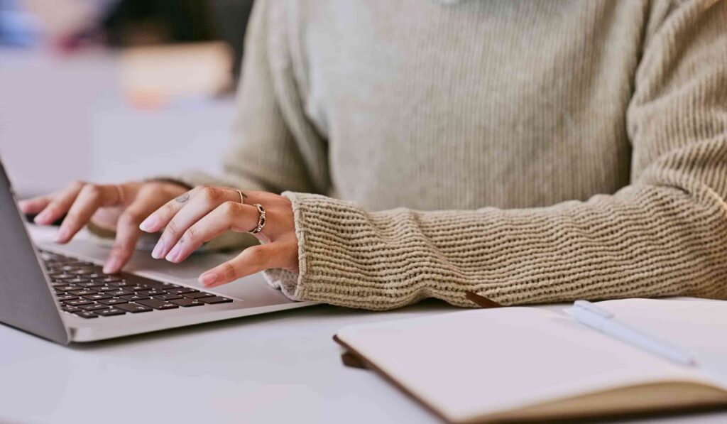 A person typing on a laptop with a notebook next to them.