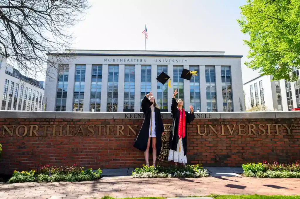 Two Northeastern graduates through their caps in the air while posing for photos at Krentzman Quad