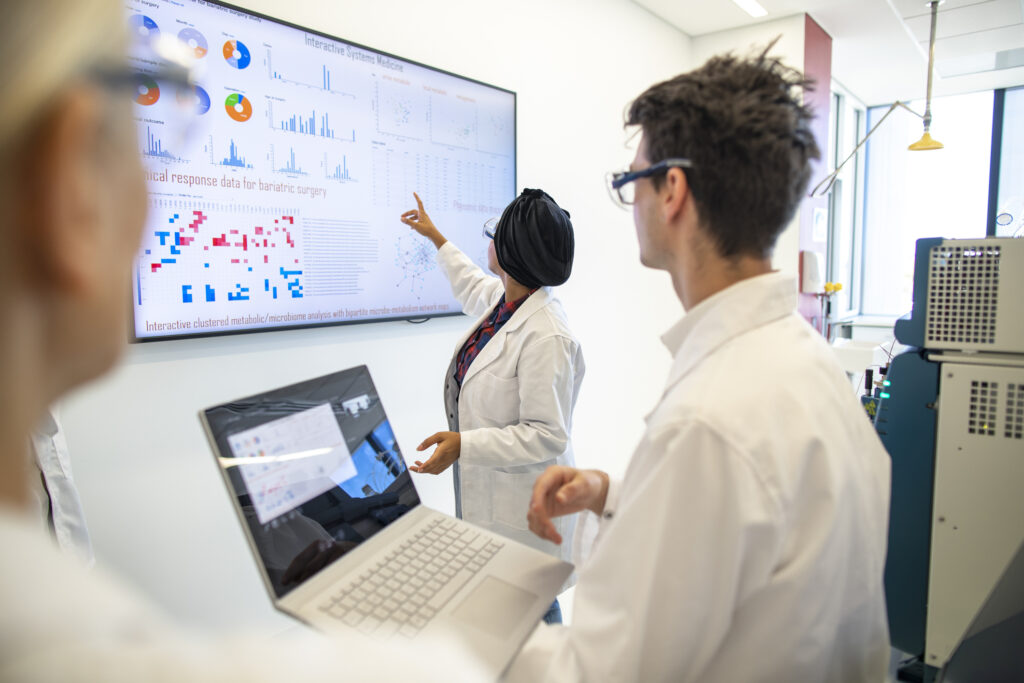 Professional team of medical scientists in a laboratory gathered around a wall monitor discussing the impact of one or more genetic variants on the Phenome providing a snapshot of a participant's health, lifestyle and environmental exposures.