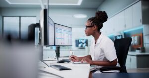 A person taking notes in front of a dual screen monitor.