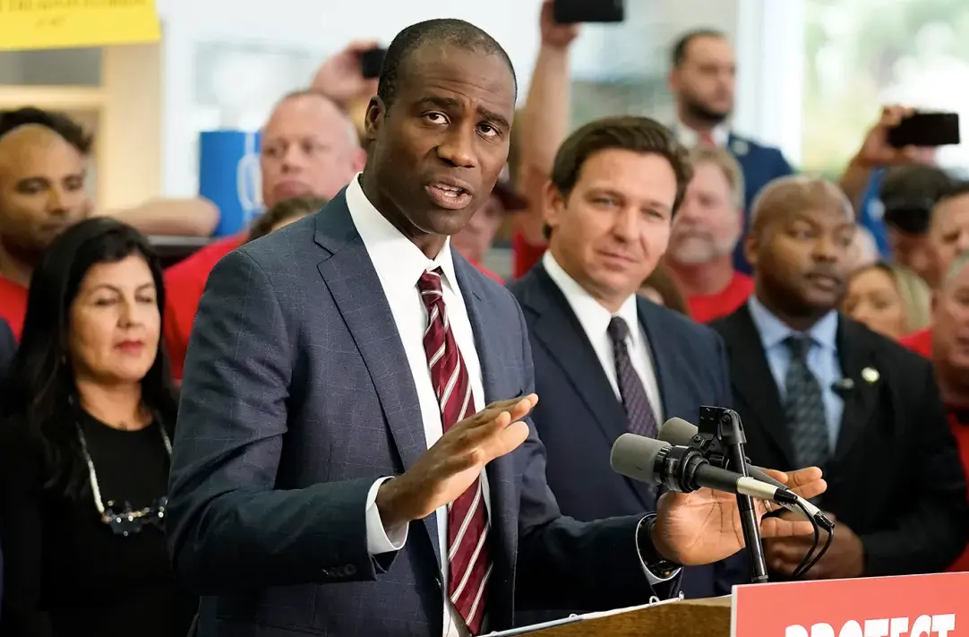 Florida Surgeon General Dr. Joseph Ladapo standing at a podium next to Governor Ron DeSantis amid a press conference announcing the state's plan to phase out childhood vaccination mandates